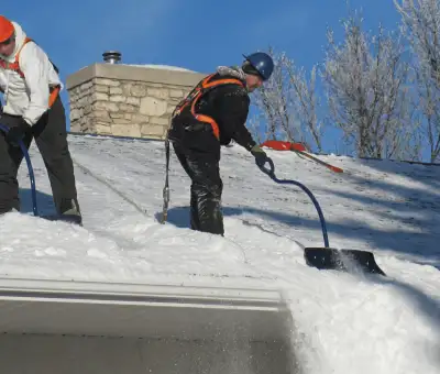 Professional technicians performing roof snow removal with safety harnesses and shovels in Erie, PA